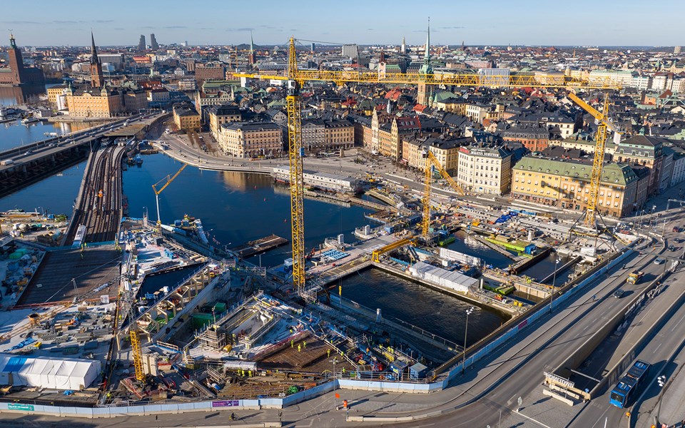 Drönarvy över arbetsområdet vid Slussen. På vattnet mellan Södermalm och Gamla stan pågår byggarbeten. Inne på arbetsområdet finns fem gula lyftkranar.